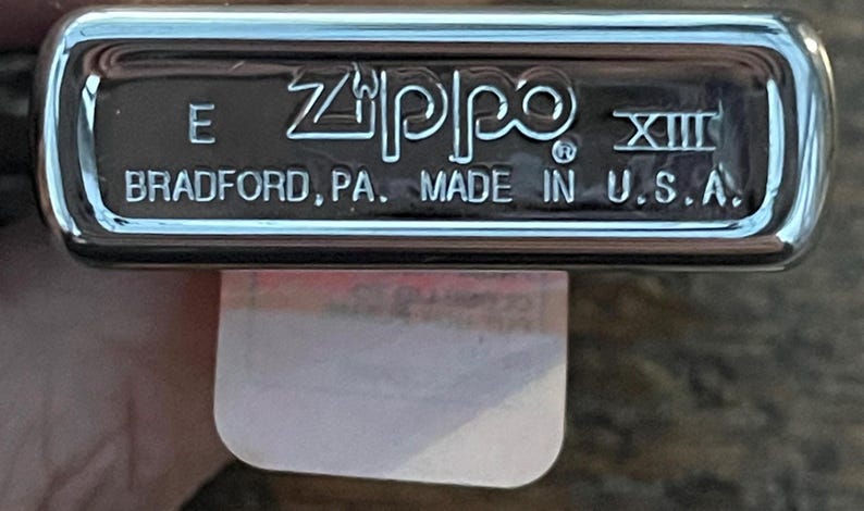 May include: A chrome-finished Zippo lighter base with the brand name and "BRADFORD, PA. MADE IN U.S.A." printed in white. The lighter has a rectangular shape with rounded edges. The Roman numeral XIII is also visible.