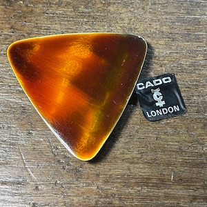 May include: A triangular, orange and yellow enamel dish with a silver rim. The dish has a label that reads "CADO LONDON" with a stylized "C" logo.