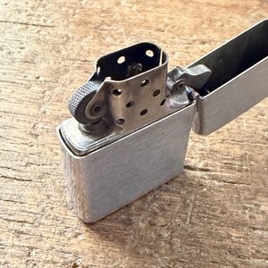 May include: A silver-colored, open lighter with a textured roller and a hinged lid. The lighter is made of metal and has a brushed finish. The lighter is resting on a wooden surface.