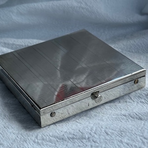 May include: A silver metal box with a textured lid and a small latch. The box is closed and sitting on a white fabric surface.