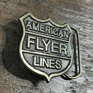 May include: A vintage brass belt buckle with the words "AMERICAN FLYER LINES" embossed on a shield-shaped design. The buckle has a tarnished, antique finish, and a metal loop for the belt.