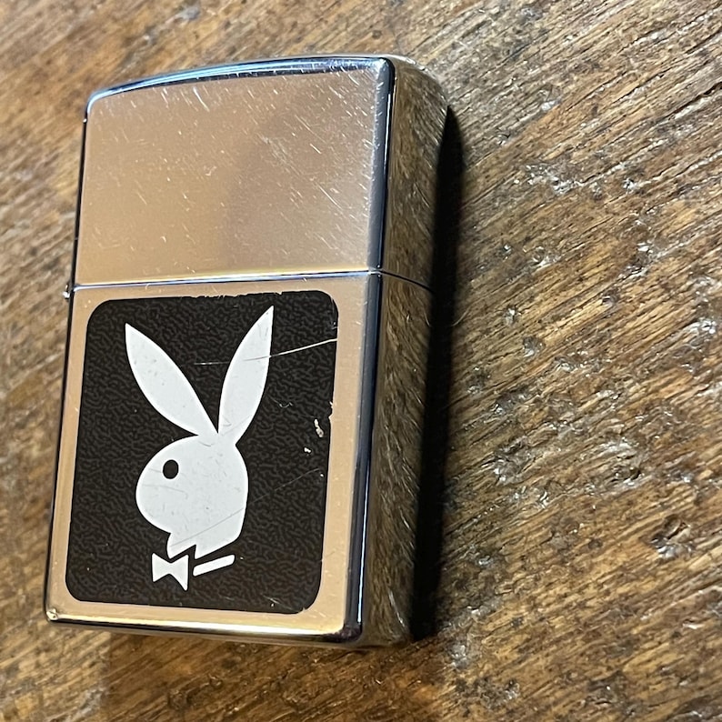 K&ouml;nnte beinhalten: Ein silberfarbenes Metallfeuerzeug mit einem schwarzen rechteckigen Emblem mit einem wei&szlig;en Hasenlogo. Das Feuerzeug hat eine rechteckige Form und eine gl&auml;nzende, reflektierende Oberfl&auml;che. Das Hasenlogo ist ein klassisches Design.