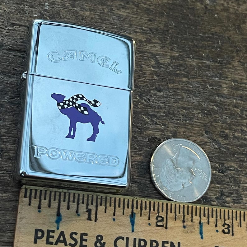 May include: A chrome-plated lighter with the word "CAMEL" at the top and "POWERED" at the bottom. The lighter features a purple camel with a checkered scarf. A quarter and a ruler are next to the lighter.