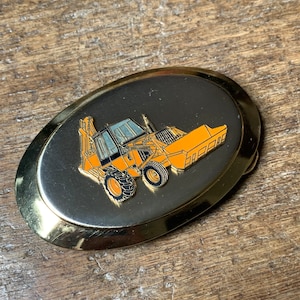 May include: A gold-tone belt buckle with a black background and a yellow and black image of a construction vehicle.