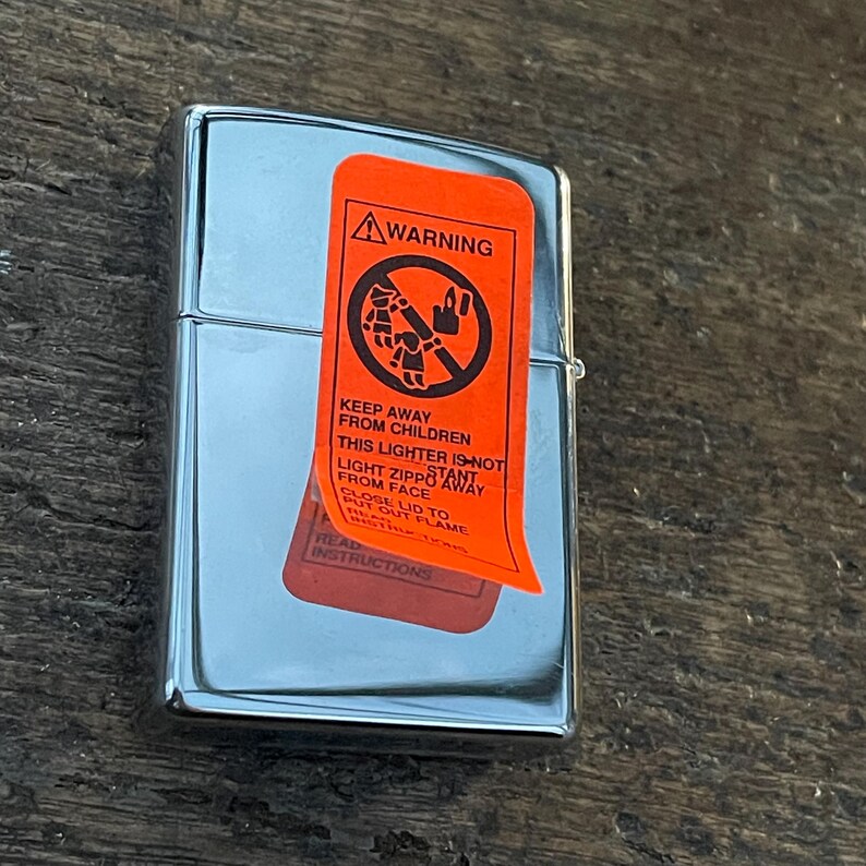 May include: A shiny, chrome-colored lighter with an orange warning label. The label has black text that reads "WARNING" and "KEEP AWAY FROM CHILDREN". The lighter is closed and appears to be new.
