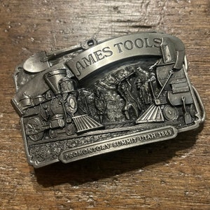 May include: Silver belt buckle featuring a detailed depiction of a steam locomotive with the text "James Tools Promontory Summit Utah 1869".
