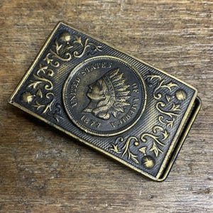 May include: A rectangular brass belt buckle with an embossed design. The center features an 1877 Indian Head cent. The buckle is framed by ornate scrollwork and floral accents. The buckle has a dark, aged finish.