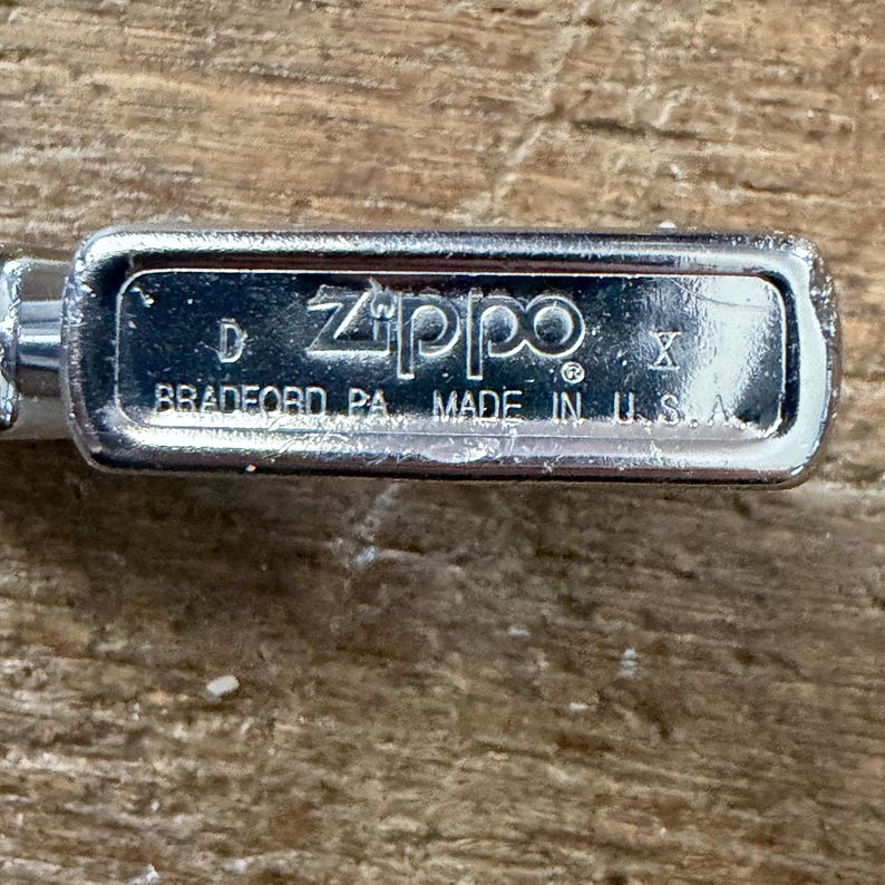 May include: Close-up of a silver Zippo lighter base. The text "Zippo" is prominently displayed, along with "BRADFORD PA. MADE IN U.S.A." on the metal surface. The lighter is set against a wooden background.