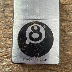 May include: A silver-colored metal lighter with a black and white eight-ball graphic. The lighter has a brushed metal finish and the text "&copy; 1993 A.A.D.L.P." is printed at the bottom. The lighter is rectangular.