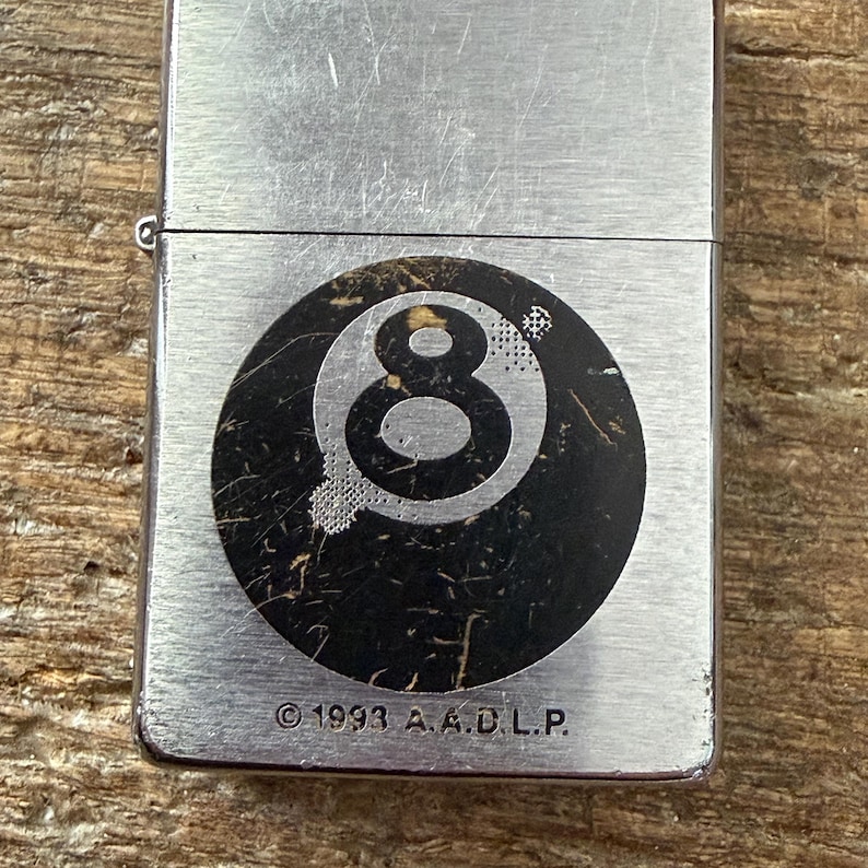 May include: A silver-colored metal lighter with a black and white eight-ball graphic. The lighter has a brushed metal finish and the text "&copy; 1993 A.A.D.L.P." is printed at the bottom. The lighter is rectangular.