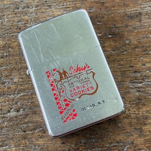 May include: A rectangular silver-tone metal lighter featuring a graphic design with red and black text that reads Icky's ORIGINAL OLD-FASHIONED VARIETY COOKIES and OLEAN, N.Y. The surface shows light scratches and a vertical red patterned border.