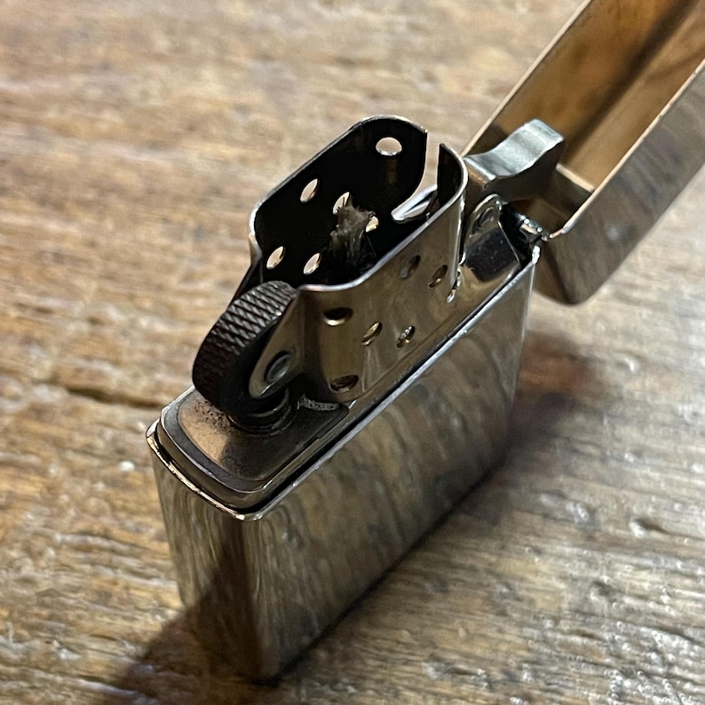 K&ouml;nnte beinhalten: Ein gl&auml;nzendes, silberfarbenes Metallfeuerzeug mit ge&ouml;ffnetem Deckel. Das Feuerzeug hat ein strukturiertes Rad zum Funken und einen kleinen Docht, der im Inneren sichtbar ist. Der Hintergrund ist eine Holzoberfl&auml;che.