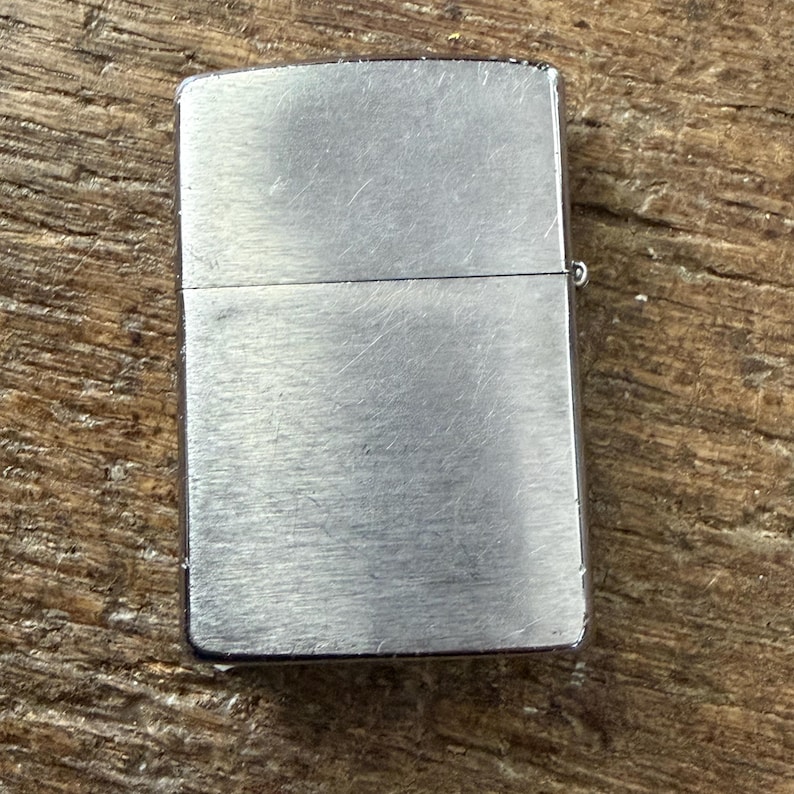 May include: A silver-colored, rectangular lighter with a hinged lid. The lighter has a brushed metal finish and a small hinge on the side. The lighter is closed and sits on a wooden surface.