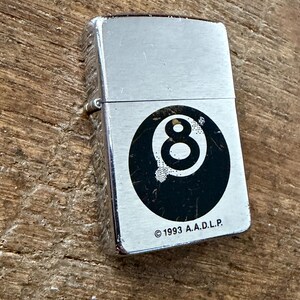 May include: A silver-colored lighter with a black and white 8-ball design. The lighter has a rectangular shape and a hinged lid. The text "&copy; 1993 A.A.D.L.P." is printed below the 8-ball graphic.