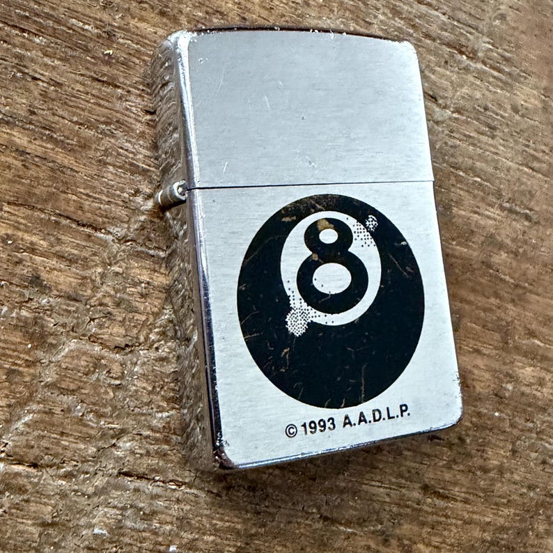 May include: A silver-colored lighter with a black and white 8-ball design. The lighter has a rectangular shape and a hinged lid. The text "&copy; 1993 A.A.D.L.P." is printed below the 8-ball graphic.