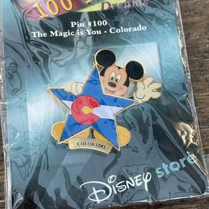 May include: A Disney pin featuring Mickey Mouse inside a blue star with the Colorado state flag design. The pin is gold-colored with the text "COLORADO" at the bottom. The packaging reads "100 YEARS of Dreams" and "Disney store".