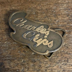 May include: Vintage brass belt buckle with the words "Charles Chips" in a cursive font. The buckle has a unique shape and a tarnished finish, giving it a retro look. The buckle is a classic accessory.