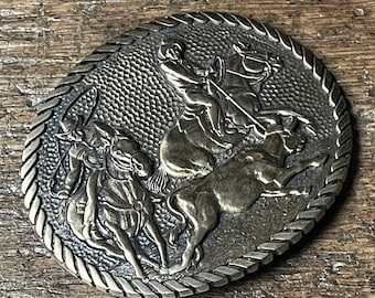 May include: A silver-toned belt buckle with a detailed design of three cowboys on horseback. The buckle features a rope design around the edge.