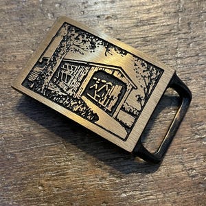 May include: A brass belt buckle with a detailed engraved image of a wooden cabin in the woods. The buckle is rectangular with a black metal loop on the side.