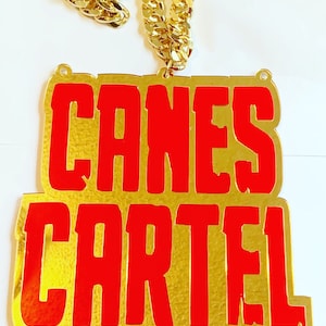 Könnte beinhalten: Goldkette mit einem großen Anhänger, auf dem die Worte "CANES CARTEL" in roten Buchstaben auf goldenem Hintergrund stehen.