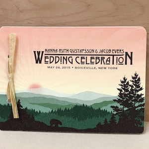 May include: A rectangular card with a pink and cream gradient sky over green layered mountains and a dark forest silhouette featuring a small bear. The text reads Hanna-Ruth Gustafsson & Jacob Evers Wedding Celebration May 24, 2015 Boiceville, New York. A raffia tie is attached to the left side.