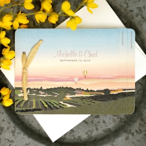 May include: A rectangular wedding invitation card featuring a landscape illustration of a vineyard at sunset with two hot air balloons, text reading Michelle & Chad September 14, 2018, and a light tan raffia ribbon tied on the left side.