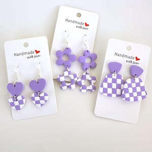 May include: Three pairs of handmade earrings on white cards. The earrings feature purple and white checkered patterns and solid purple shapes, including hearts, flowers, and geometric designs. Each card has the text "Handmade with love" and a small red heart.