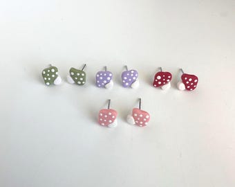 May include: A collection of mushroom-shaped stud earrings in various colors, including green, purple, red, and pink. Each earring features a white polka dot pattern and a white base. The earrings are displayed on a white surface.