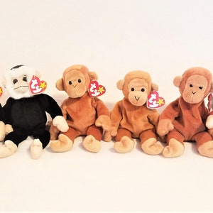 May include: A collection of six plush monkey beanie babies. The monkeys are arranged in a row from left to right. The first two monkeys are black with white faces. The next two monkeys are brown. The fifth monkey is a darker brown. The last monkey is red. All of the monkeys have white tags with red stars.