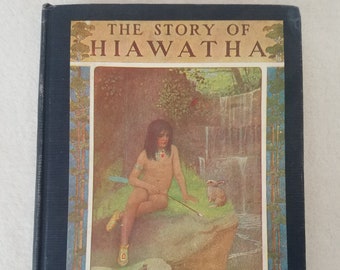 Könnte beinhalten: Der Einband eines Buches mit dem Titel "The Story of Hiawatha". Der Einband zeigt eine farbenfrohe Illustration eines jungen indianischen Jungen, der auf einem Felsen an einem Wasserfall sitzt. Er hält einen Stock und ein kleines Tier sitzt neben ihm.
