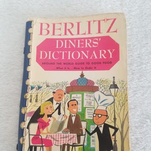 Könnte beinhalten: Ein Vintage-Buchcover von Berlitz Diners' Dictionary mit einer Cartoon-Illustration eines Kochs, eines Kellners und einer Frau, die an einem Tisch in einem Pariser Café sitzen. Der Buchtitel lautet "Berlitz Diners' Dictionary" und der Untertitel lautet "Around the World Guide to Good Food... What It Is... How to Order It".