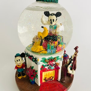 May include: A musical snow globe featuring Mickey Mouse, Pluto and Goofy. Mickey is standing on a pile of presents, Pluto is sitting on the presents, and Goofy is playing a bass. The snow globe is decorated with a Christmas wreath, a fireplace and a poinsettia.