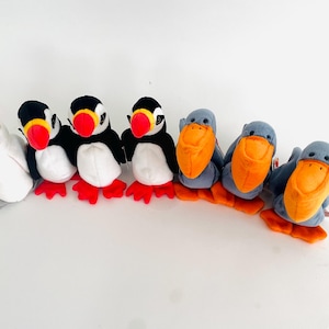 May include: A collection of seven plush stuffed animals, including a white bird with a pink tuft, three black and white puffins, and three gray birds with orange beaks.