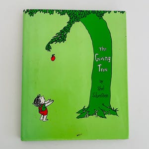 May include: The cover of a green book titled "The Giving Tree" by Shel Silverstein. The cover features a green tree with a red apple on a branch and a small boy with his arms outstretched.