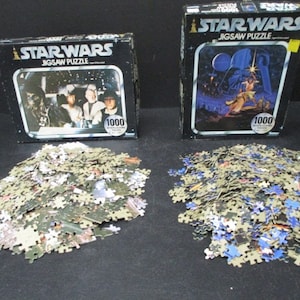 Puede incluir: Dos rompecabezas de Star Wars sin abrir de 1000 piezas. La caja de la izquierda presenta una escena de la película con Luke Skywalker, Han Solo y Chewbacca. La caja de la derecha presenta una escena con Darth Vader y la princesa Leia.