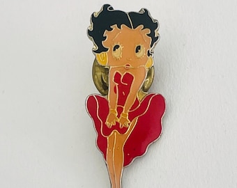 Op de afbeelding: Een rode en gouden emaille pin met een cartoonfiguur van Betty Boop die een rode jurk draagt met een gouden riem en gouden oorbellen.