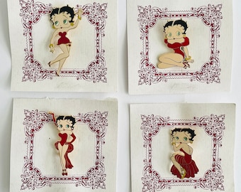 Op de afbeelding: Vier Betty Boop emaille pins in verschillende poses, elk geplaatst in een decoratief bordeauxrood kader op een getextureerde papieren achtergrond. De pins tonen Betty Boop in een rode jurk met zwart haar en gouden accenten.