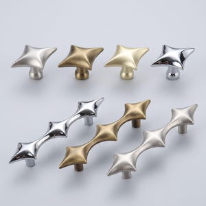 May include: A collection of four square-shaped cabinet knobs and three elongated cabinet pulls, each featuring a four-pointed star design. The hardware is finished in various metallic tones, including polished chrome, brushed gold, and matte silver.