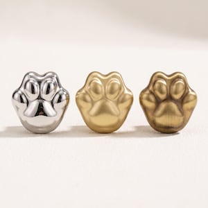 Könnte beinhalten: Drei Möbelknöpfe aus Metall in Form von Tierpfoten. Der erste Knopf ist aus poliertem Silber, der zweite aus mattem Gold und der dritte hat ein gebürstetes Antik-Messing-Finish. Jeder Knopf zeigt ein zentrales Ballenpolster und vier Zehen.