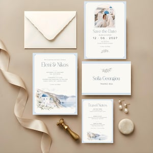 Könnte beinhalten: Ein Hochzeitspapeterie-Set mit blauen Rändern, bestehend aus Save-the-Date-Karte, Einladung, Antwortkarte und Reisehinweisen. Mit Aquarellillustrationen von Santorin und dem Text Eleni & Nikos, 12 06 2027.