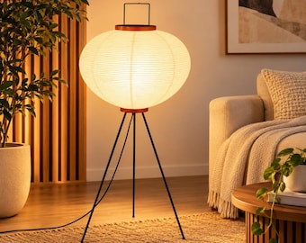 May include: A floor lamp featuring a large, round, off-white paper lantern shade with horizontal ribbing, topped with a thin black metal handle. The shade is supported by a slender, three-legged black metal tripod base standing on the floor.