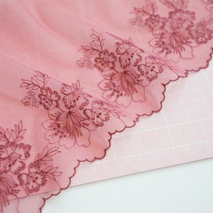 Op de afbeelding: Close-up van een delicate roze kanten stof met geborduurde bloempatronen in een donkerdere roze tint. Het kant heeft een geschulpte rand en het bloemmotief bevat gedetailleerde bloemen en bladeren.