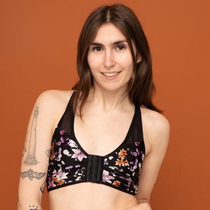 May include: Black floral print sports bra with a hook-and-eye closure. The bra features a halter neckline with mesh detailing and a vibrant pattern of pink, purple, and orange flowers. The background is a solid orange color.