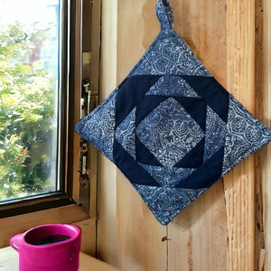 May include: A blue and white quilted pot holder with a geometric pattern. The pot holder is hanging on a wooden wall.