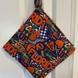 May include: A square, quilted pot holder with a loop for hanging. The fabric features a repeating pattern of orange, blue, and green elements, including the word "FLORIDA", alligators, and sports-related graphics.