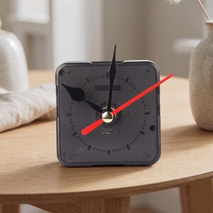 May include: A square-shaped analog clock with a dark gray face and a dotted circular minute track. The clock features two black spade-shaped hands for hours and minutes, and a thin, bright red second hand centered on a gold-toned cap.