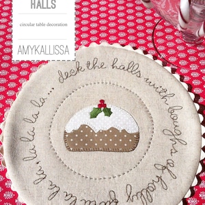 Puede incluir: Una decoración de mesa circular beige con el texto "deck the halls with boughs of holly" bordado alrededor del borde. En el centro hay una imagen bordada en marrón y blanco de un pudín navideño con hojas de acebo verdes en la parte superior.