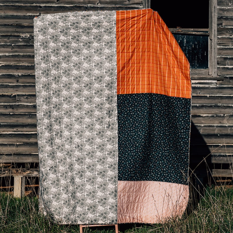 K&ouml;nnte beinhalten: Eine Patchwork-Decke mit vier verschiedenen Stoffmustern. Die Decke besteht aus einem wei&szlig;en Stoff mit einem schwarzen Blumenmuster, einem orangefarbenen Stoff mit wei&szlig;en Streifen, einem dunkelblauen Stoff mit einem wei&szlig;en Blumenmuster und einem rosa Stoff mit einem wei&szlig;en Steppmuster.