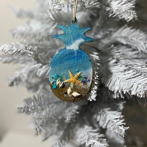 May include: A blue and white pineapple-shaped Christmas ornament with a beach scene inside. The ornament features a starfish, seashells, and sand.