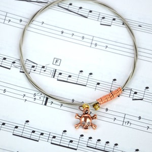 May include: A silver guitar string bracelet with a copper skull charm. The bracelet is lying on a sheet of music with musical notes.